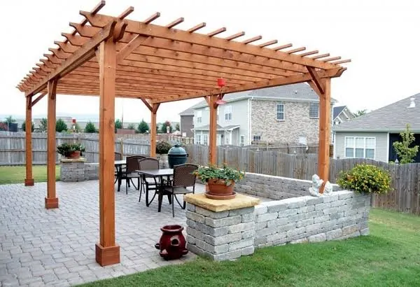 Taş döşemeli bahçe terasında ahşap pergola, altında masa ve sandalyeler, yanında taş örgülü alçak duvar ve çiçekli saksılar