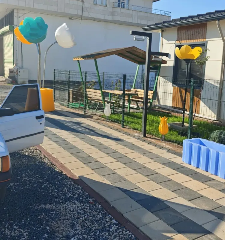 Güneşli havada kilit parke döşeli yolun kenarında renkli lale biçimli armatürler, yeşil tel çit, ahşap çatılı oturma çardağı ve arkada ahşap detaylı tek katlı çelik ev