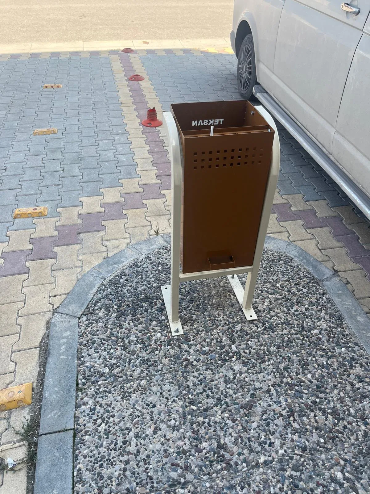 Bej boru ayaklı çerçevede duran kahverengi delikli metal çöp kutusu; çevresinde kilit parke ve çakıllı zemin