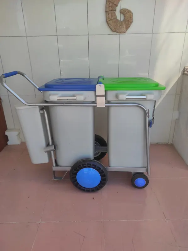 Metal çerçeveli, mavi ve yeşil kapaklı iki gri hazne taşıyan, mavi saplı itme kollu tekerlekli çöp arabası