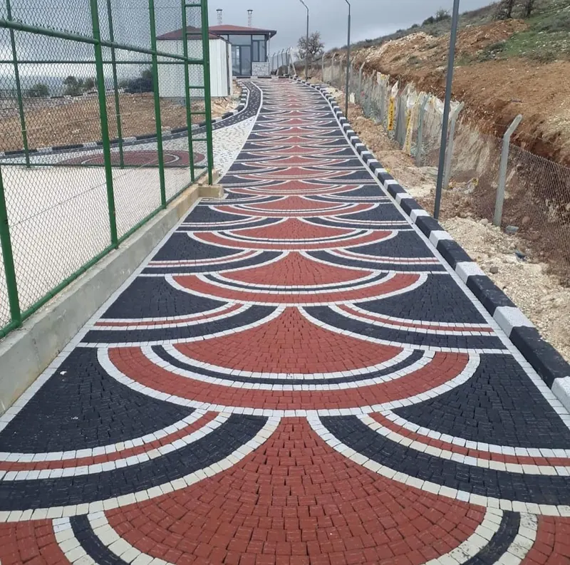 Kırmızı, siyah ve beyaz kilit parkelerle yelpaze deseni oluşturulmuş, kenarı bordürlü, tel örgülü bir sahanın yanından yukarı uzanan yaya yolu